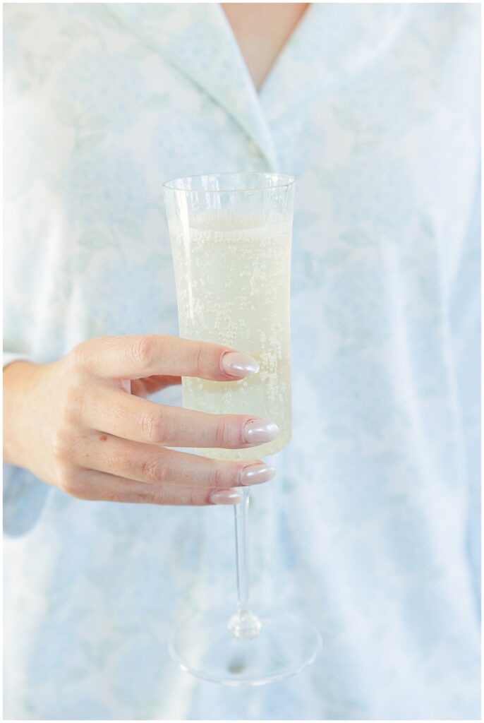 Person wearing light blue floral pajamas holding a glass of sparkling wine or champagne, part of wedding morning preparations at Nauset Beach Inn in Orleans, MA.