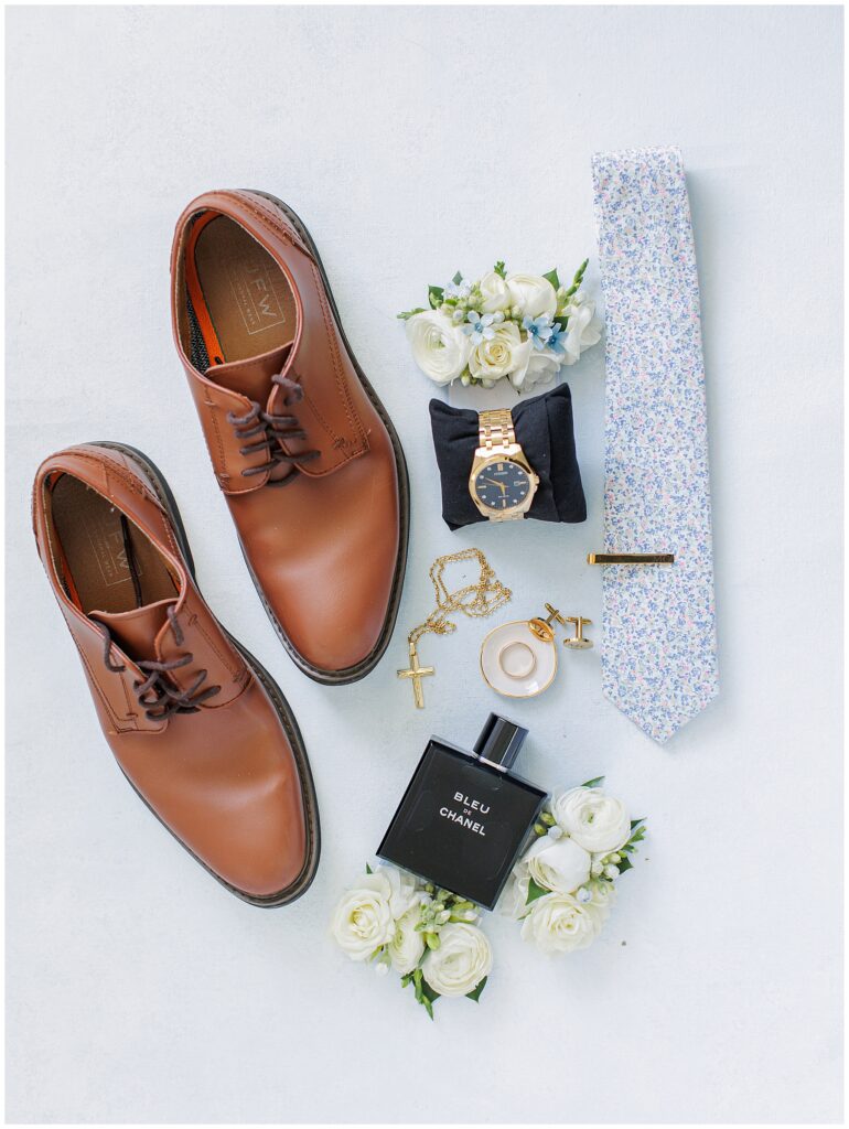 Flat lay of groom’s wedding details on a light blue surface, including brown leather dress shoes, a floral tie, gold watch, boutonniere, gold cufflinks, a cross necklace, wedding rings, and a bottle of Bleu de Chanel cologne, photographed at Nauset Beach Inn in Orleans, MA.