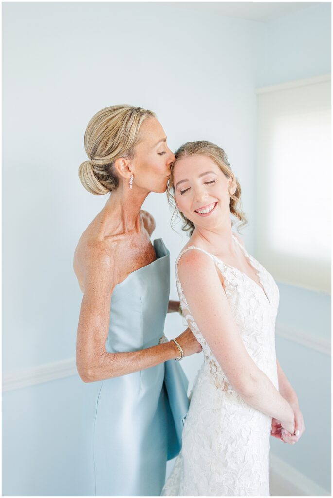 Bride smiling as an older woman kisses her on the forehead while helping her get ready, both dressed in elegant light-colored attire in a softly lit room at Nauset Beach Inn in Orleans, MA.