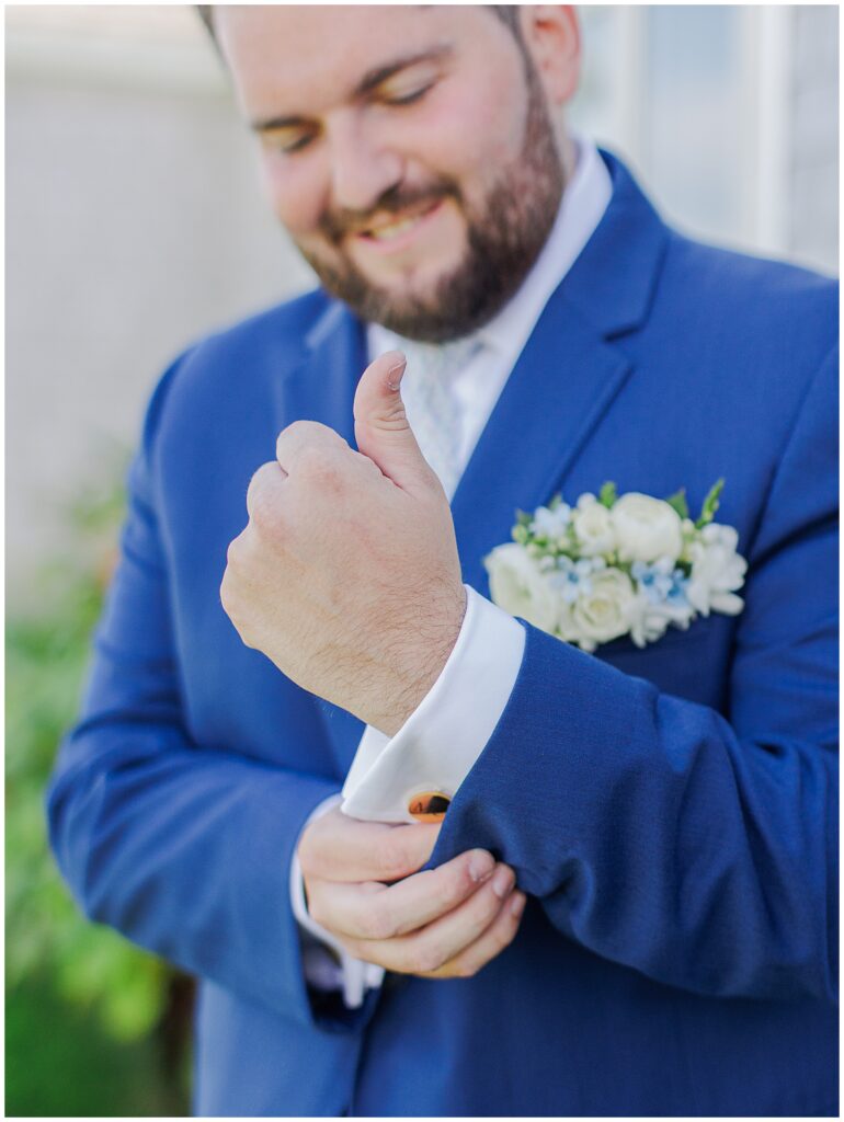 Groom in a navy suit adjusting his cufflinks, smiling outdoors with a white floral boutonniere pinned to his jacket at Nauset Beach Inn in Cape Cod.