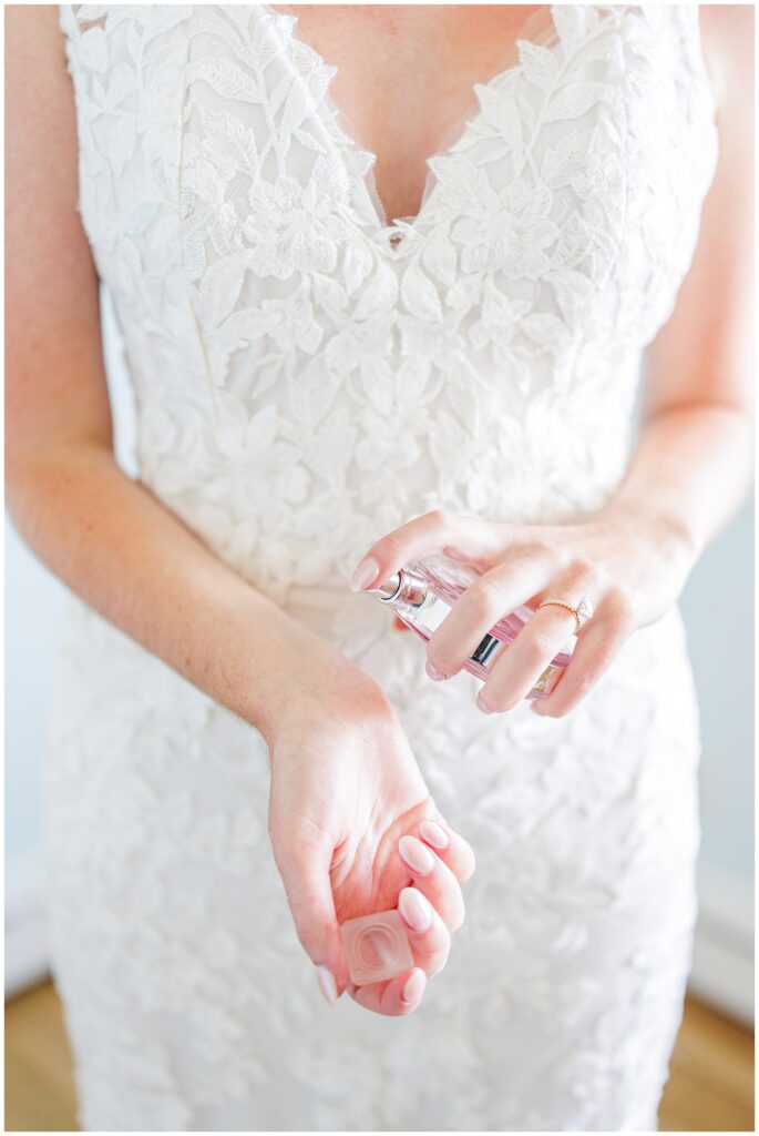 Bride in a lace wedding gown spraying perfume on her wrist while getting ready, photographed indoors with soft natural light at Nauset Beach Inn in Orleans, MA.