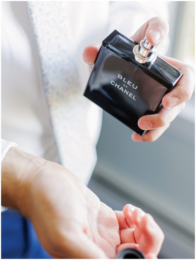 Close-up of groom’s hands applying Bleu de Chanel cologne to his wrist while wearing a floral tie and white dress shirt, taken during wedding preparations at Nauset Beach Inn in Cape Cod.