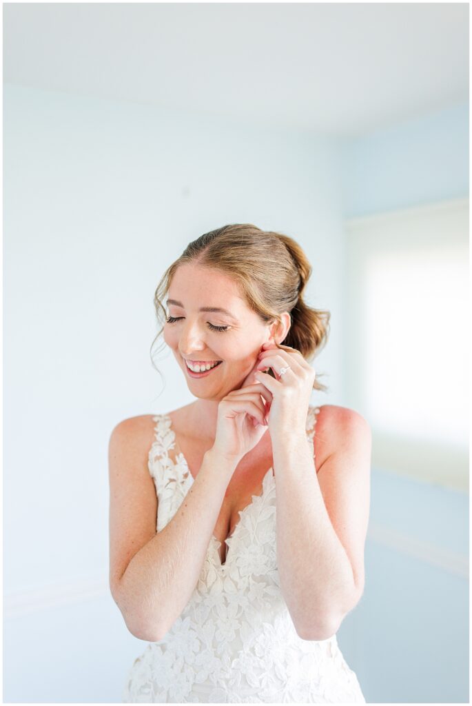 Bride smiling while putting on earrings, wearing a lace gown and styled hair, captured during her wedding morning preparations at Nauset Beach Inn in Orleans, MA.