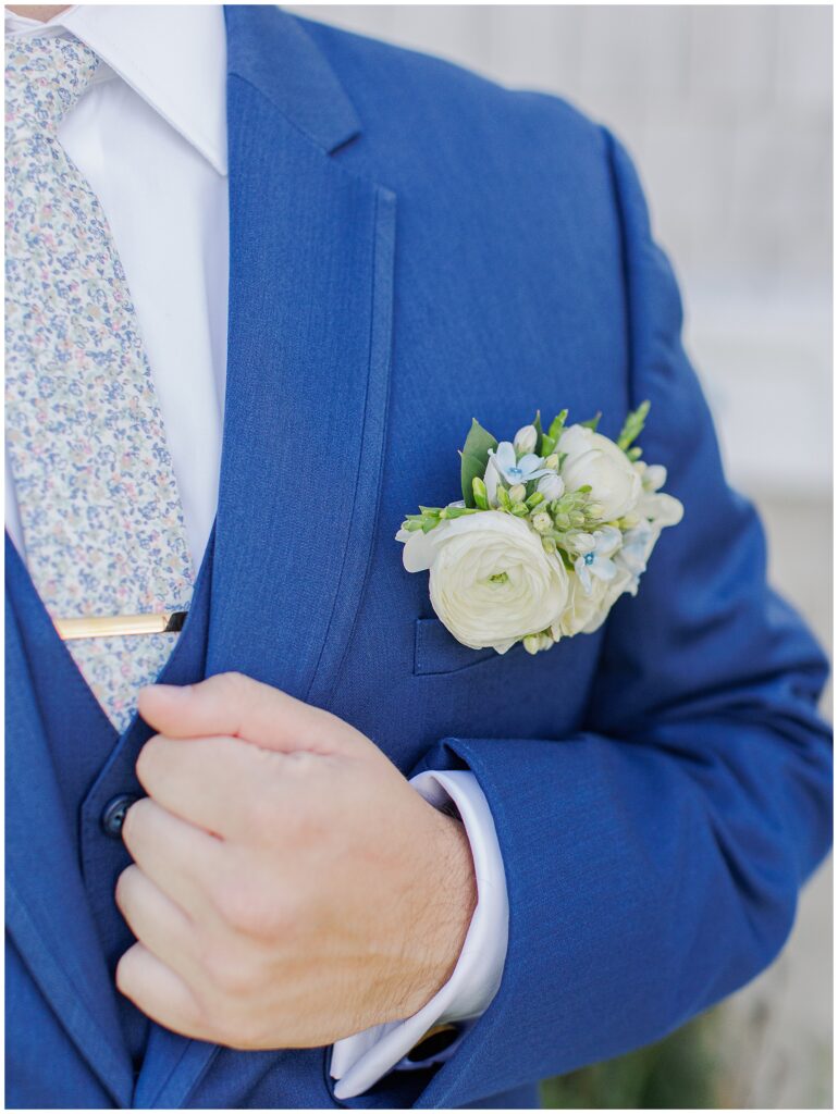 Close-up of groom’s navy blue suit showing a floral tie, white shirt, and white boutonniere with blue accents, taken outside Nauset Beach Inn in Cape Cod.