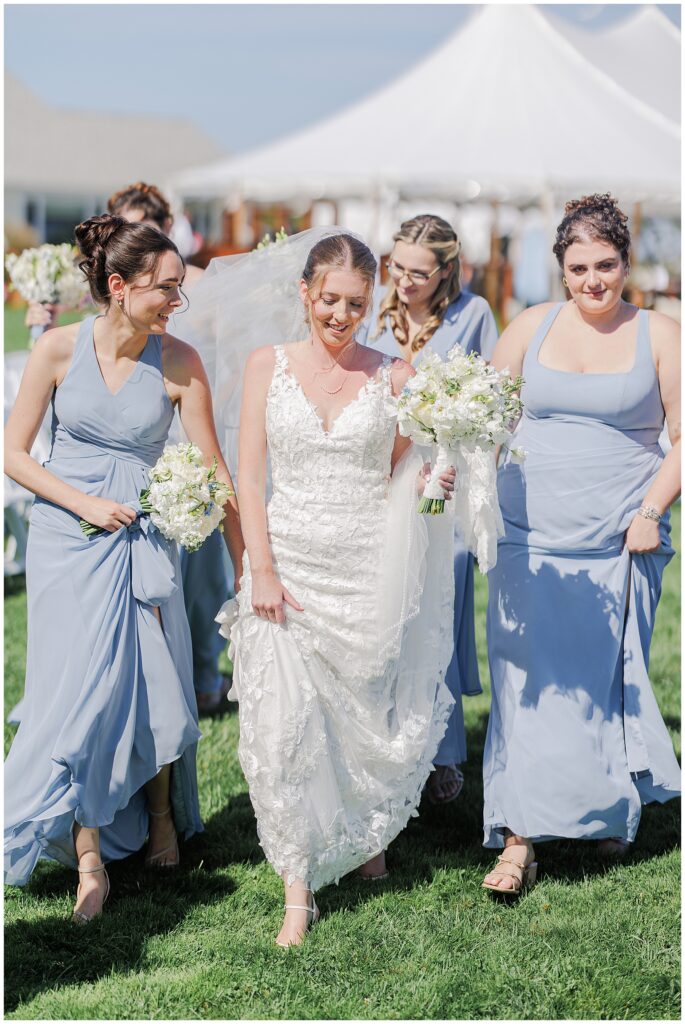 Bride walking across the grass with bridesmaids dressed in light blue gowns, all holding white flower bouquets, with a white tent visible in the background at Nauset Beach Inn in Orleans, MA.