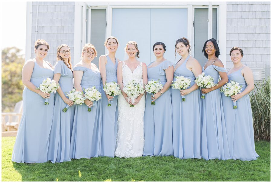 Bride standing with her bridesmaids in matching light blue dresses, each holding white bouquets, posing in front of a shingled building at Nauset Beach Inn in Cape Cod.