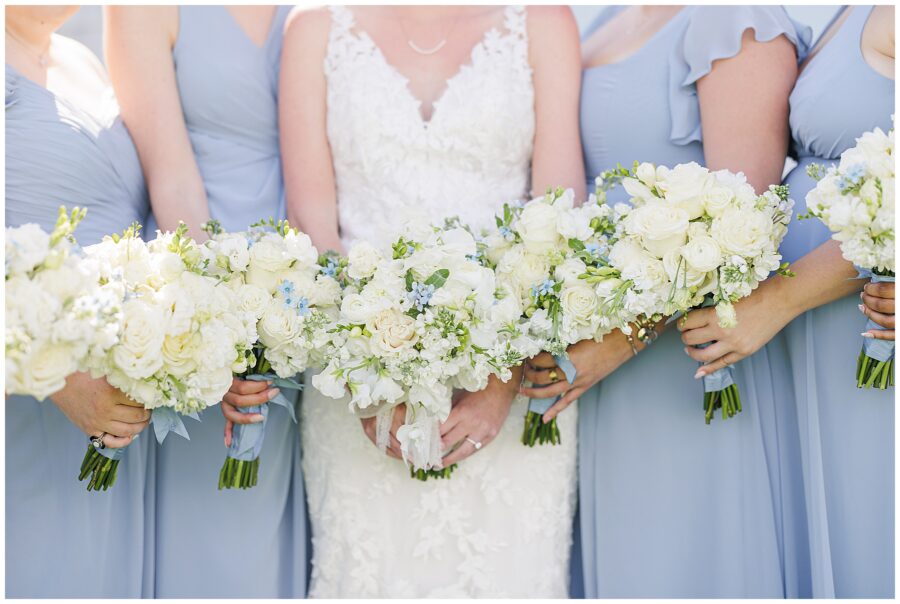 Close-up of the bride and bridesmaids holding white and cream floral bouquets with small blue accents, their light blue dresses and lace wedding gown coordinated for a coastal wedding at Nauset Beach Inn in Orleans, MA.