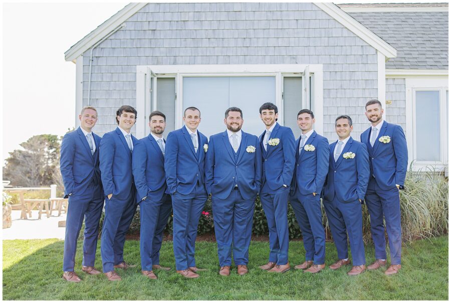Groom with his groomsmen dressed in matching navy blue suits, posing in front of a gray shingled building at Nauset Beach Inn in Cape Cod.