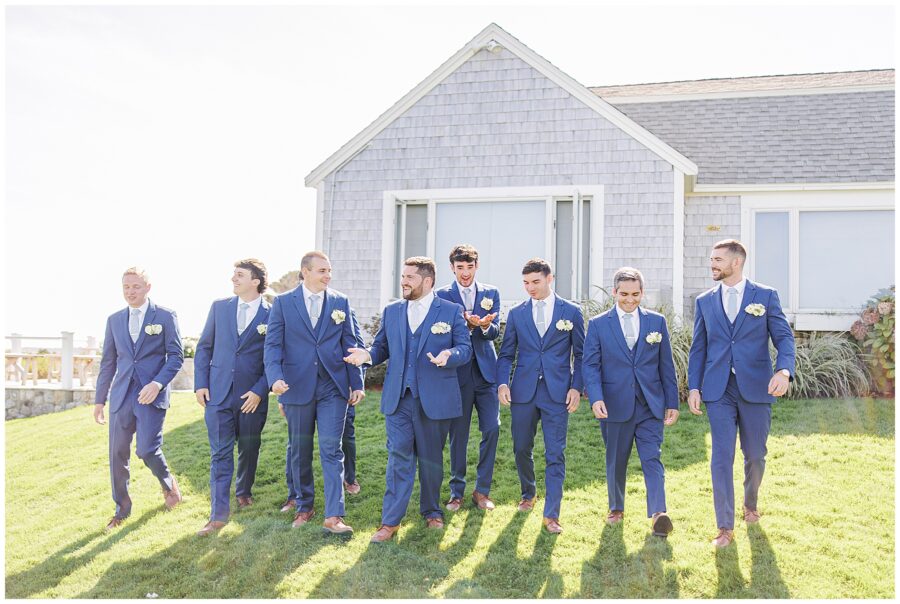 Groomsmen in navy suits walking and laughing together on the lawn outside Nauset Beach Inn in Orleans, MA, with coastal cottages visible in the background.