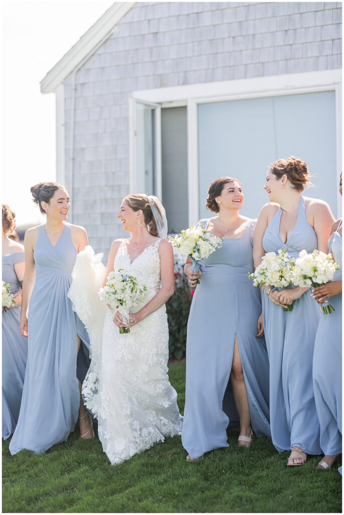 Bride walking and smiling with her bridesmaids dressed in light blue gowns, holding white floral bouquets outside the gray shingled cottages at Nauset Beach Inn in Cape Cod.