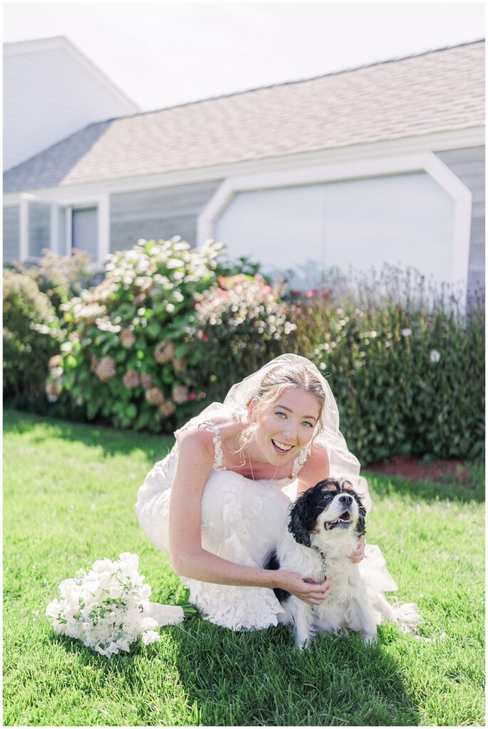 Bride crouching on the grass and smiling beside a small black and white dog, holding her white floral bouquet at Nauset Beach Inn in Orleans, MA.