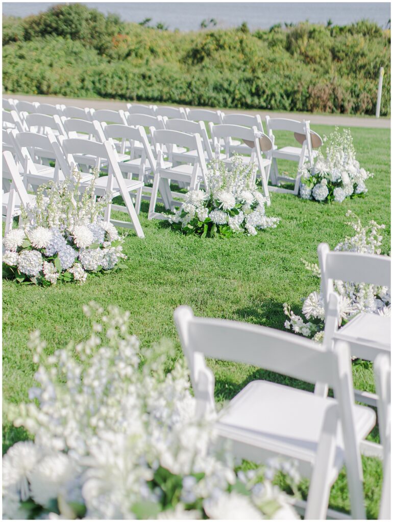 Close-up of white floral arrangements and rows of white ceremony chairs on the green lawn at Nauset Beach Inn in Orleans, MA, with ocean views in the background.