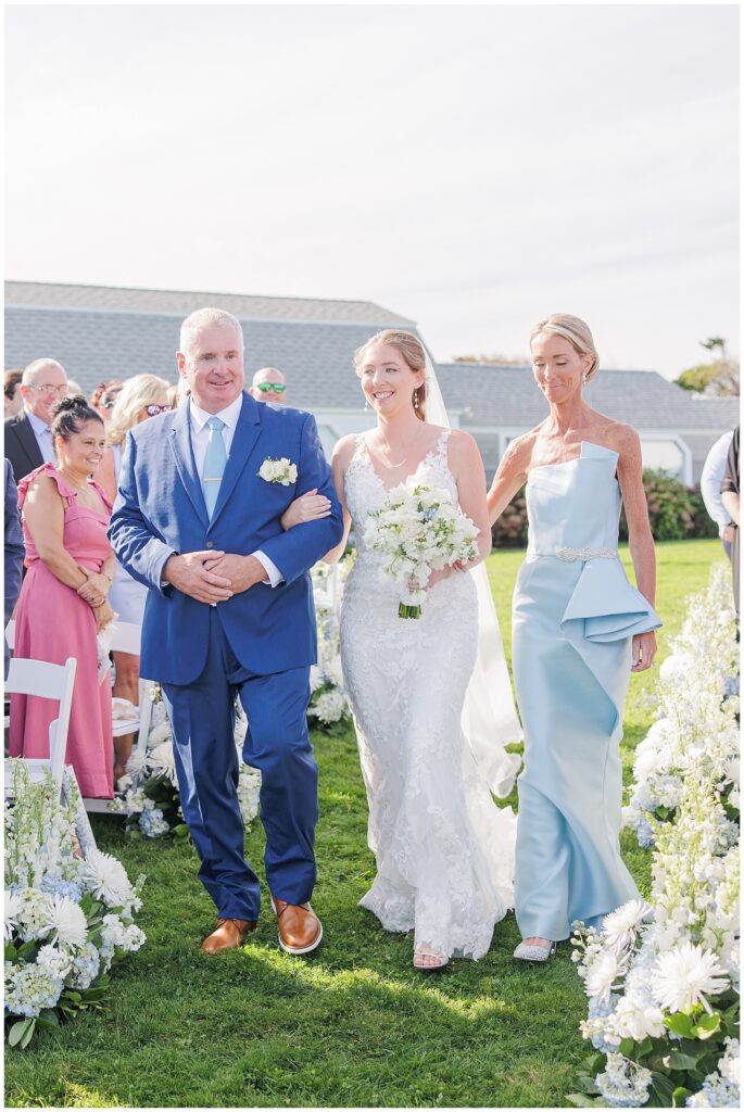 Bride walking down the aisle between two people dressed in blue, surrounded by white floral arrangements and wedding guests on the lawn at Nauset Beach Inn in Cape Cod.
