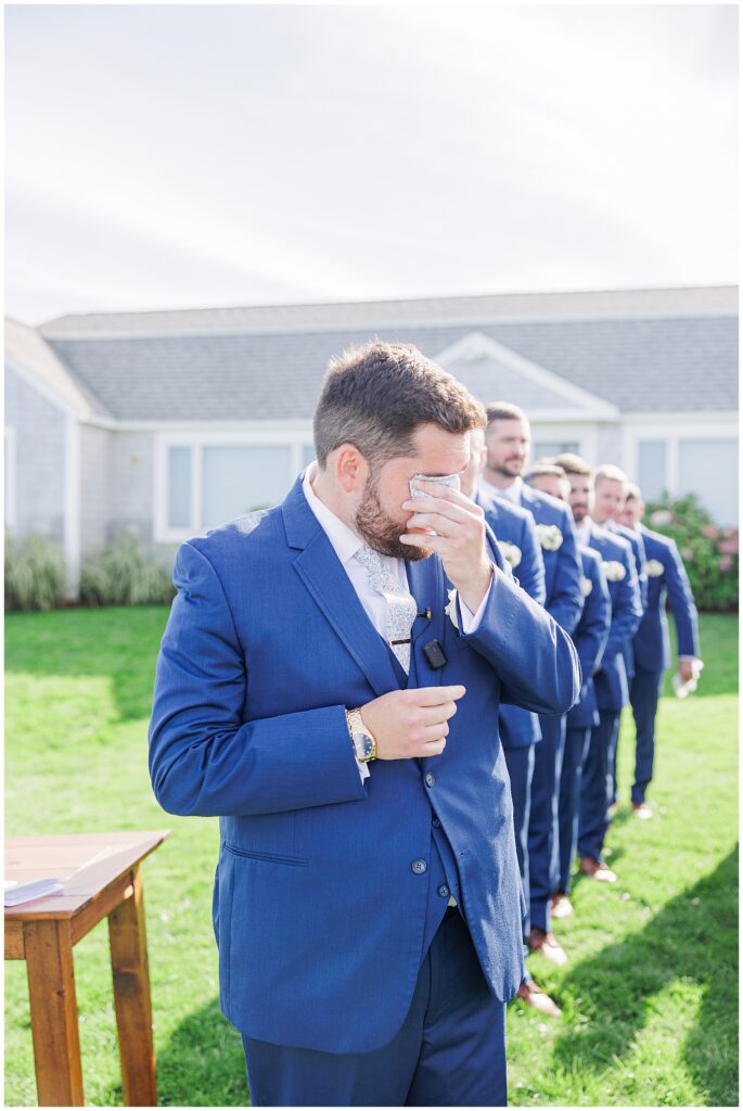 Groom in a navy suit wiping away tears as he stands with his groomsmen during the outdoor ceremony at Nauset Beach Inn in Orleans, MA.