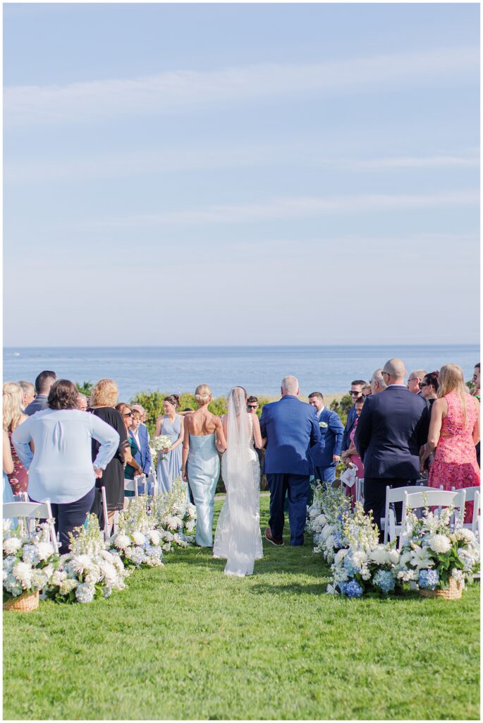 Bride walking down the aisle between two people toward the groom, surrounded by seated guests and white floral arrangements on the oceanfront lawn at Nauset Beach Inn in Orleans, MA.
