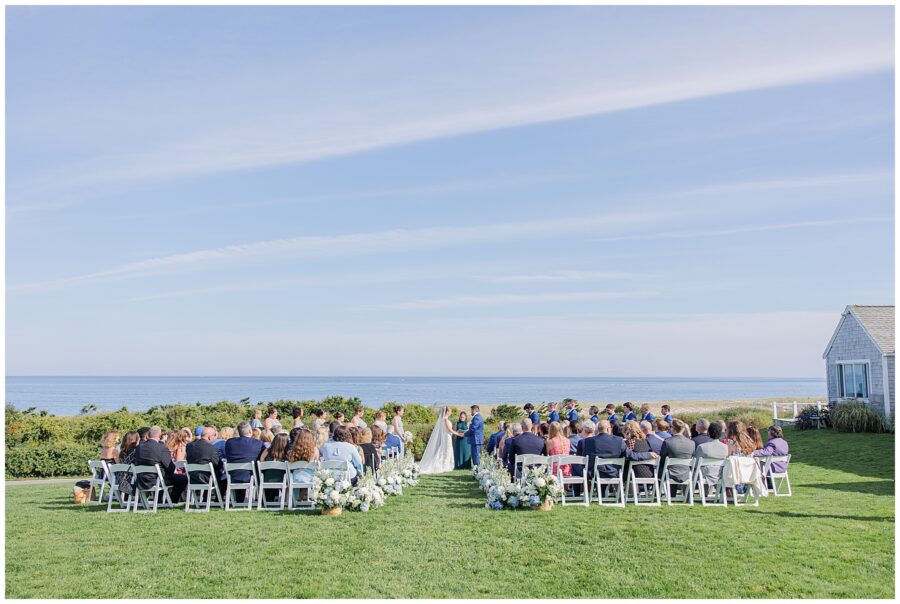 Outdoor wedding ceremony overlooking the Atlantic Ocean at Nauset Beach Inn in Cape Cod, with guests seated in white chairs and the couple standing together at the altar.