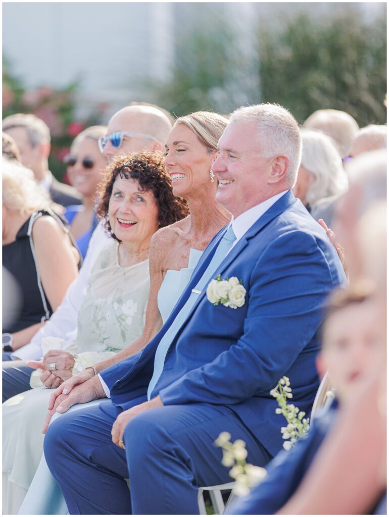 Guests seated in the front row smiling during the outdoor ceremony at Nauset Beach Inn in Orleans, MA, with the inn’s shingled cottages visible in the background.