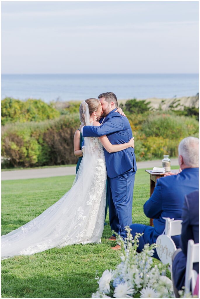 Bride and groom sharing their first kiss as a married couple during the ceremony on the lawn at Nauset Beach Inn in Cape Cod, with the ocean in the background.