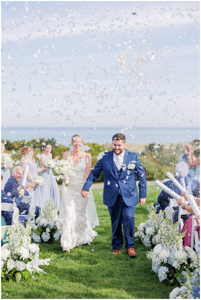 Bride and groom walking up the aisle as guests celebrate with white confetti, surrounded by floral arrangements and ocean views at Nauset Beach Inn in Orleans, MA.