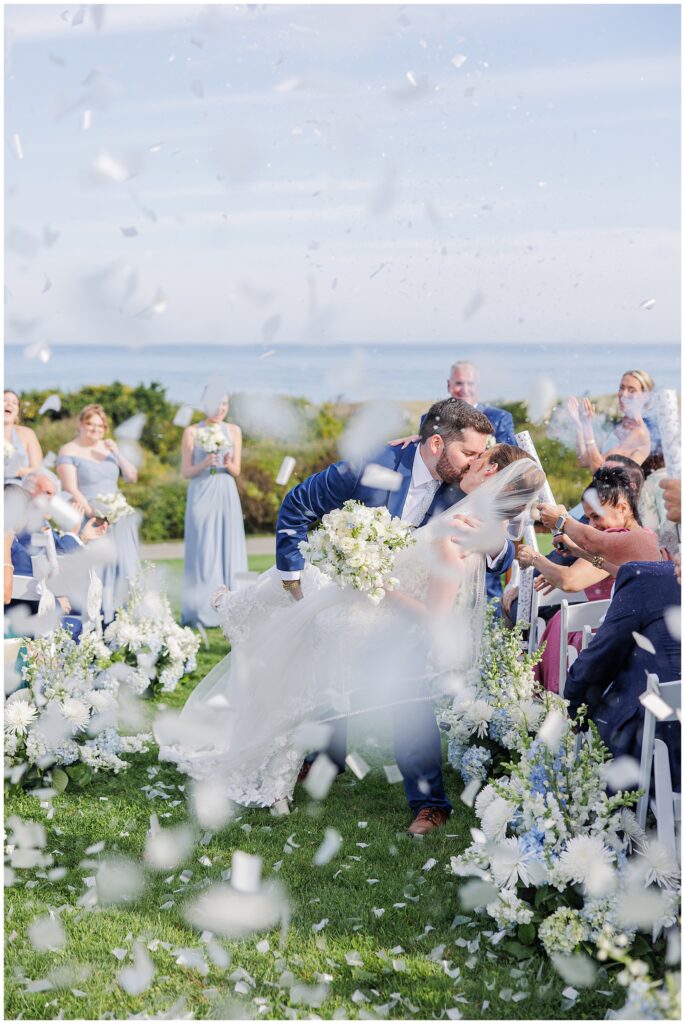 Bride and groom kissing under falling confetti after their ceremony, surrounded by cheering guests and coastal scenery at Nauset Beach Inn in Cape Cod.