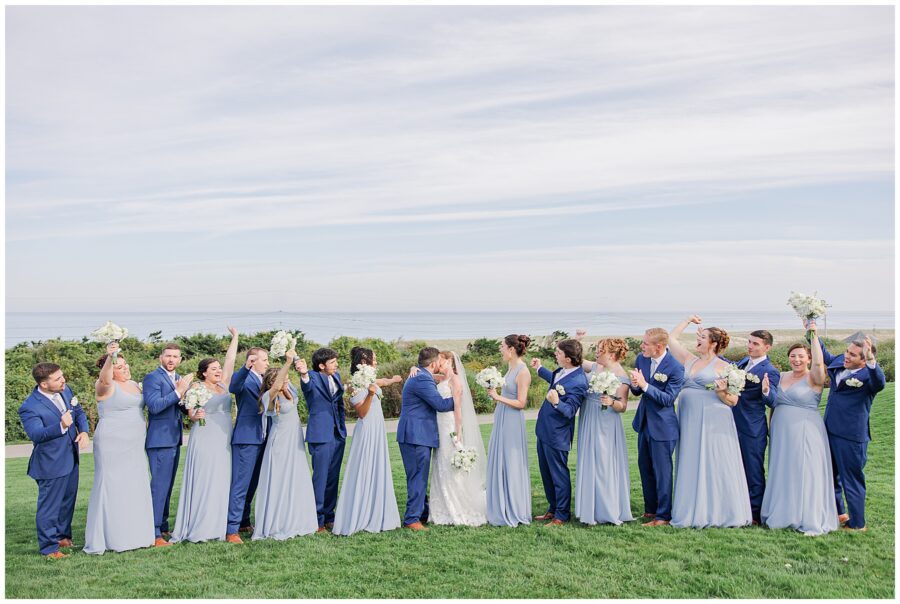 Bride and groom kissing while surrounded by their wedding party dressed in light blue gowns and navy suits, celebrating on the lawn at Nauset Beach Inn in Orleans, MA.