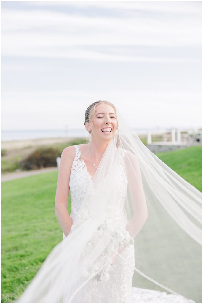 Bride smiling with her veil blowing in the breeze, standing on the green lawn with ocean views behind her at Nauset Beach Inn in Cape Cod.