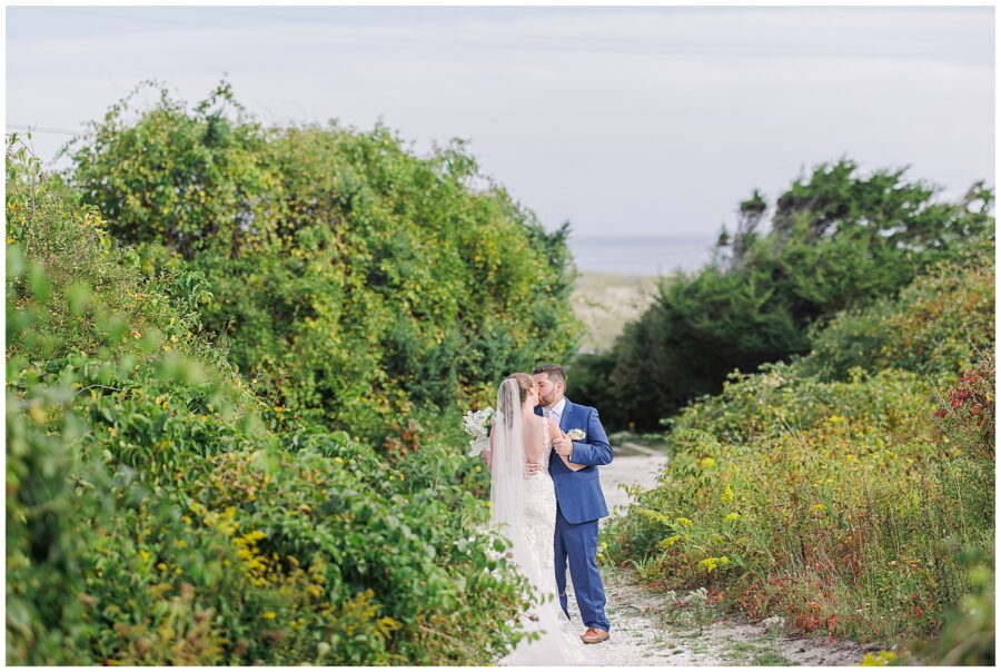 Bride and groom kissing on a sandy path surrounded by greenery, with a glimpse of the ocean in the distance near Nauset Beach Inn in Cape Cod.