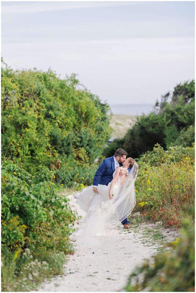 Groom dipping the bride for a kiss on a white sand path lined with greenery, captured near the ocean at Nauset Beach Inn in Orleans, MA.