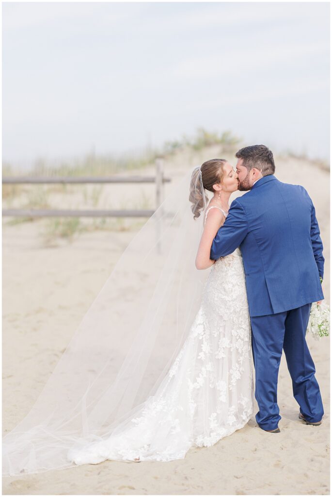 Bride and groom kissing on the sand beside a wooden fence, with beach dunes in the background at Nauset Beach Inn in Cape Cod.