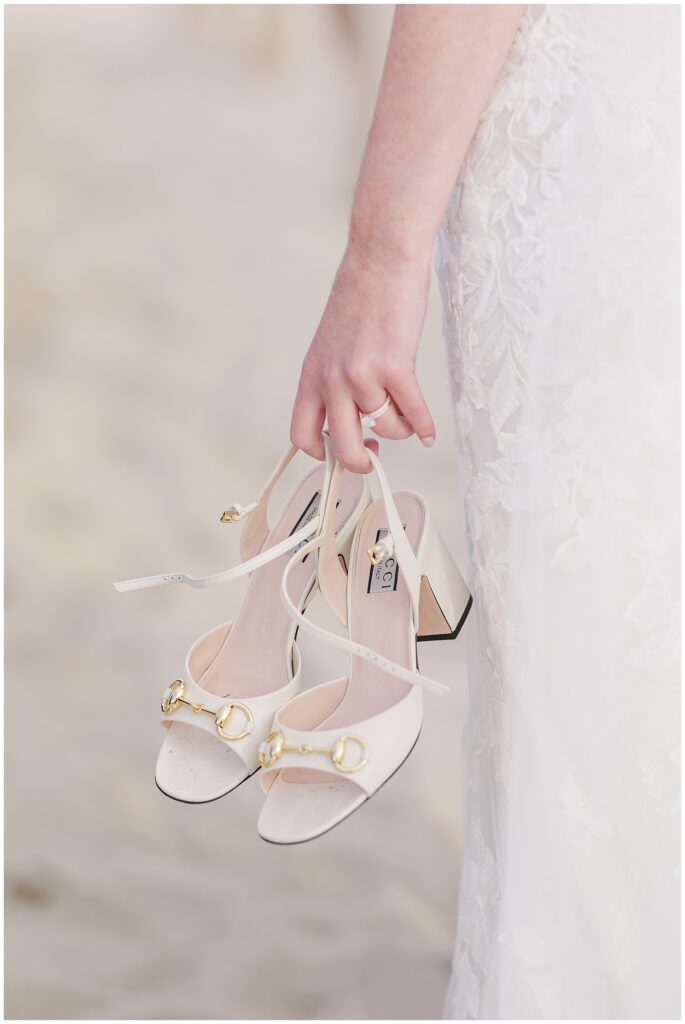 Close-up of the bride holding her white Gucci block-heeled sandals with gold buckles while standing barefoot on the sand at Nauset Beach Inn in Orleans, MA.