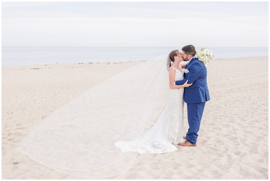 Bride and groom kissing on the beach with the bride’s long veil flowing in the breeze and the ocean in the background at Nauset Beach Inn in Cape Cod.