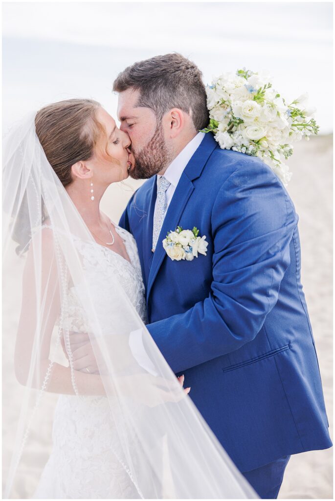 Close-up of bride and groom kissing on the sandy beach, the bride’s veil draped around them, during their Nauset Beach Inn wedding in Orleans, MA.