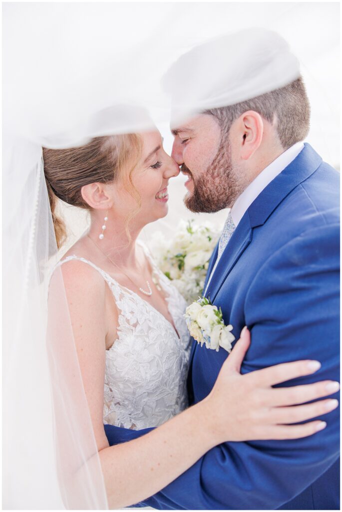 Bride and groom smiling closely under the bride’s veil, embracing and holding a white floral bouquet on the beach at Nauset Beach Inn in Cape Cod.