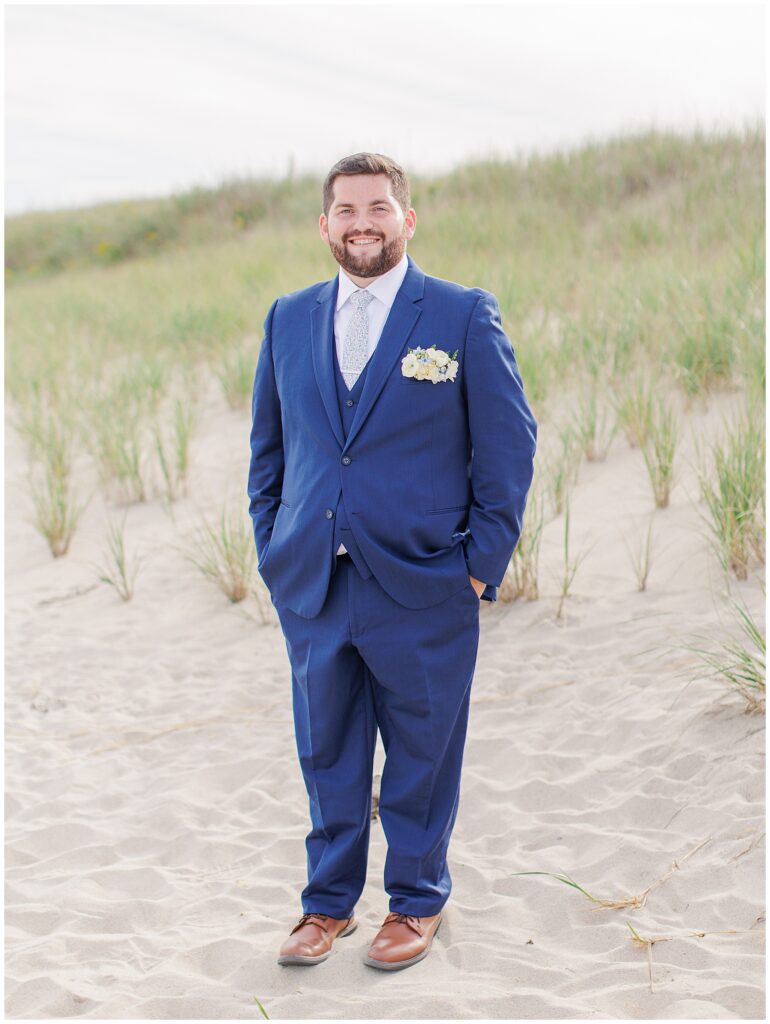 Groom standing and smiling on the sandy dunes in a navy suit with a white boutonniere, photographed at Nauset Beach Inn in Cape Cod.