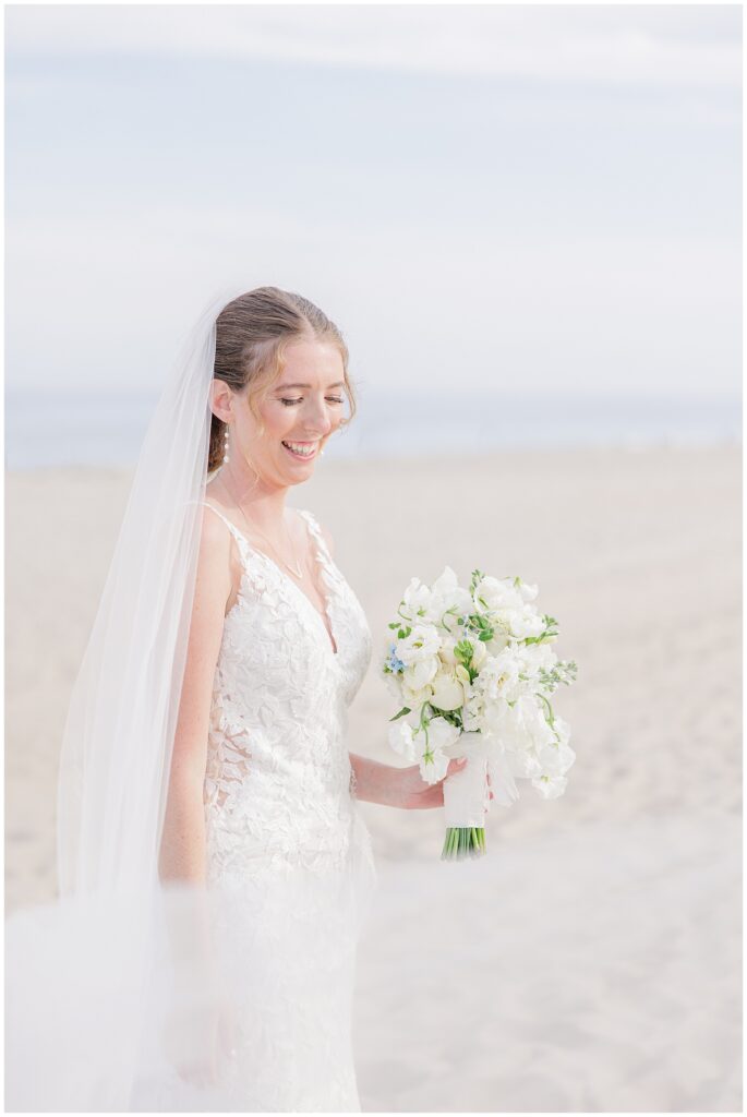 Bride smiling and holding a white floral bouquet on the sandy beach, her lace gown and long veil flowing in the coastal breeze at Nauset Beach Inn in Orleans, MA.