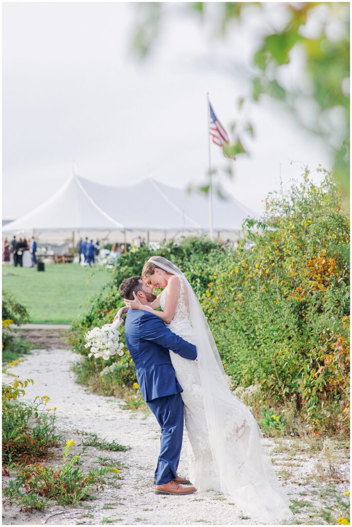 Groom lifting the bride for a kiss on a sandy path lined with green bushes, with a white reception tent and American flag visible in the background at Nauset Beach Inn in Orleans, MA.