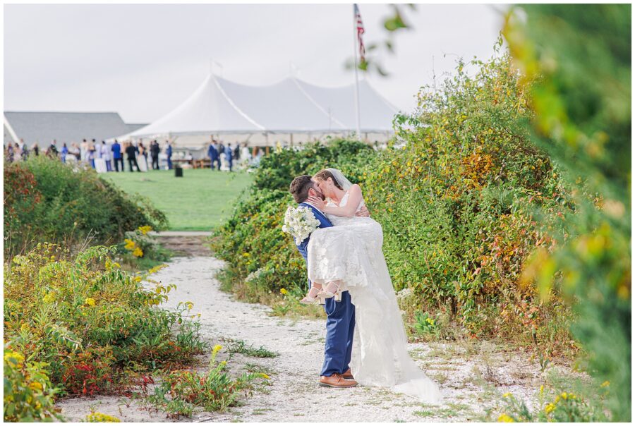 Bride and groom embracing on a white sand path surrounded by greenery, with their wedding reception tent and guests visible on the lawn at Nauset Beach Inn in Cape Cod.