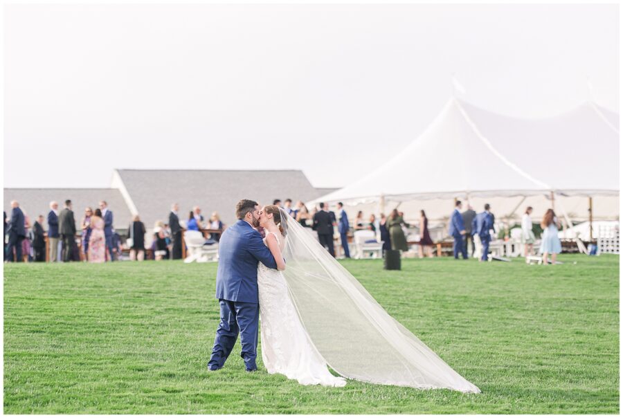 Bride and groom kissing on the green lawn in front of their white wedding tent, surrounded by guests during a celebration at Nauset Beach Inn in Orleans, MA.