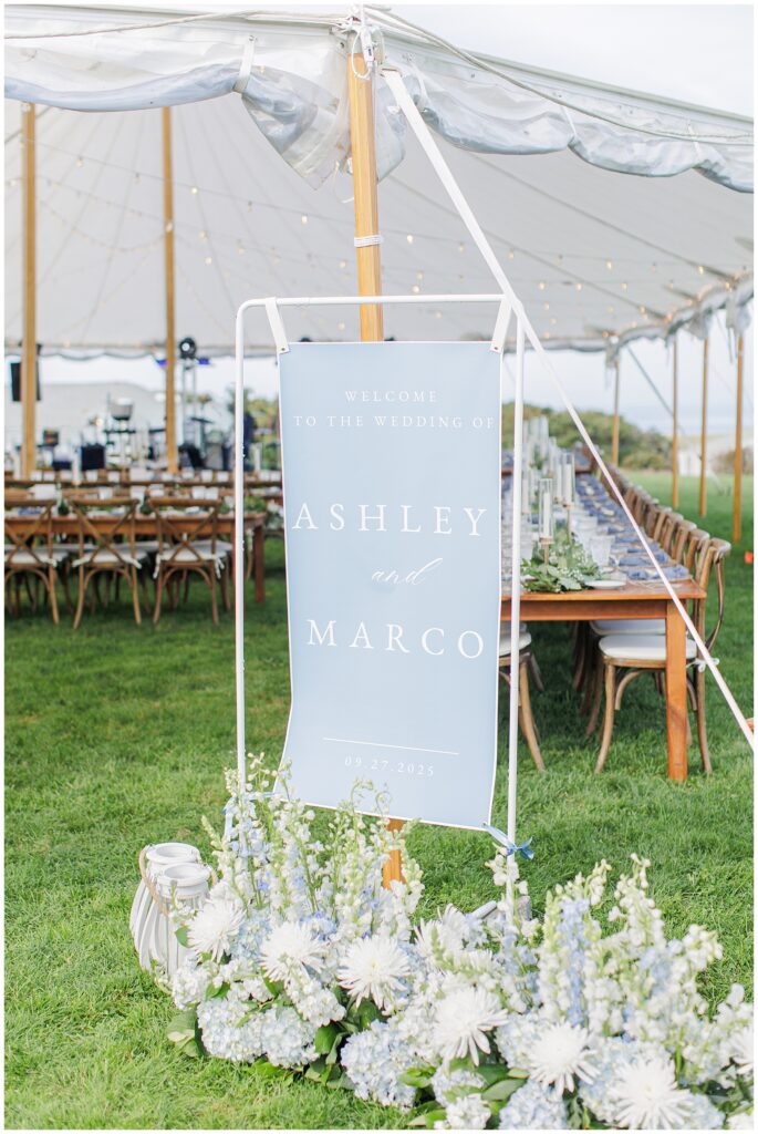 Wedding welcome sign reading “Welcome to the wedding of Ashley and Marco, 09.27.2025” displayed at the entrance of the reception tent with blue and white floral arrangements at Nauset Beach Inn in Orleans, MA.