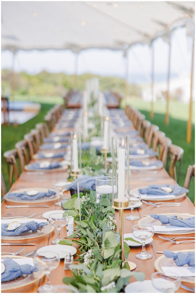 Reception table setting under a sailcloth tent featuring wooden tables, blue napkins, gold-rimmed plates, tall glass candles, and greenery runners at Nauset Beach Inn in Cape Cod.