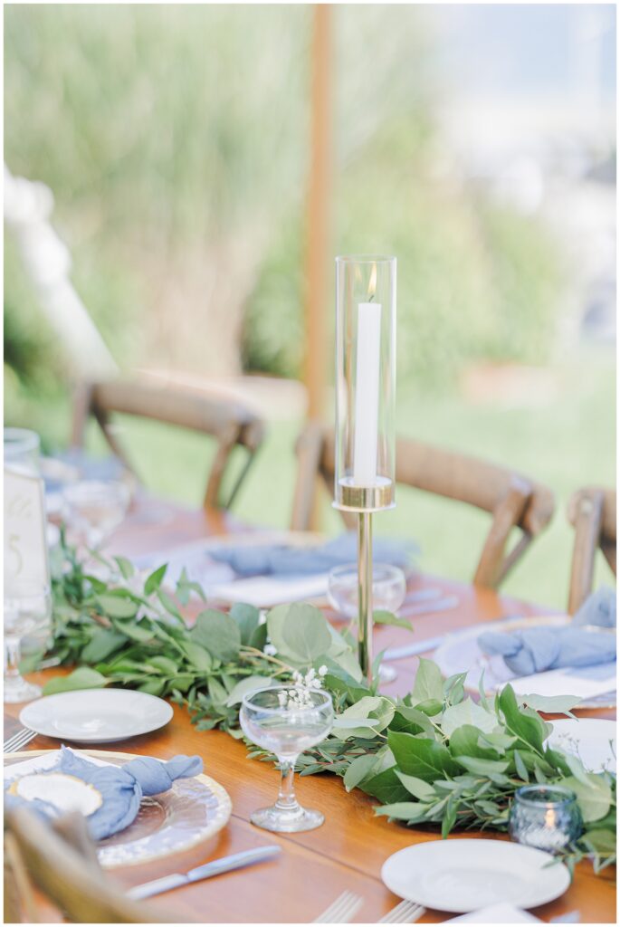 Close-up of a reception table centerpiece with greenery garlands, glass candle holders, and blue napkins arranged neatly at Nauset Beach Inn in Orleans, MA.