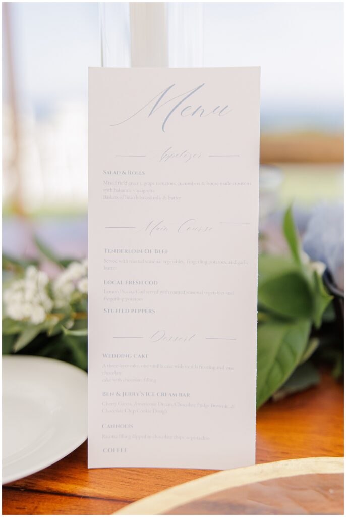 Wedding dinner menu displayed on a wooden table decorated with greenery and candles, listing salad, tenderloin of beef, fresh cod, and desserts, photographed at Nauset Beach Inn in Cape Cod.