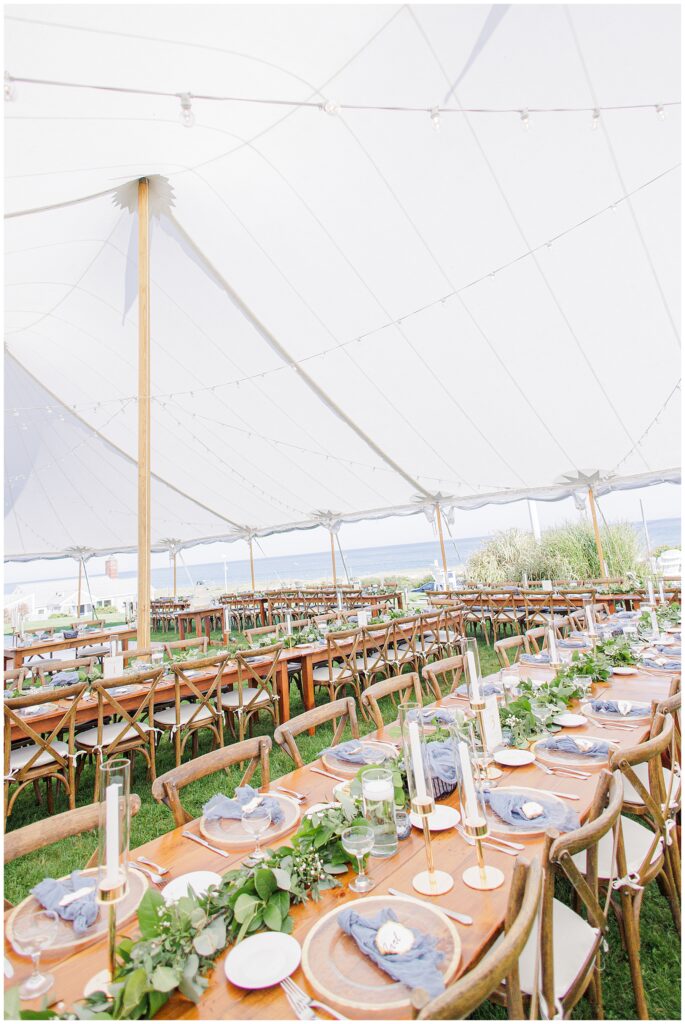 Under a white sailcloth tent overlooking the ocean, long wooden reception tables are set with blue napkins, greenery runners, and candles for a coastal wedding at Nauset Beach Inn in Orleans, MA.