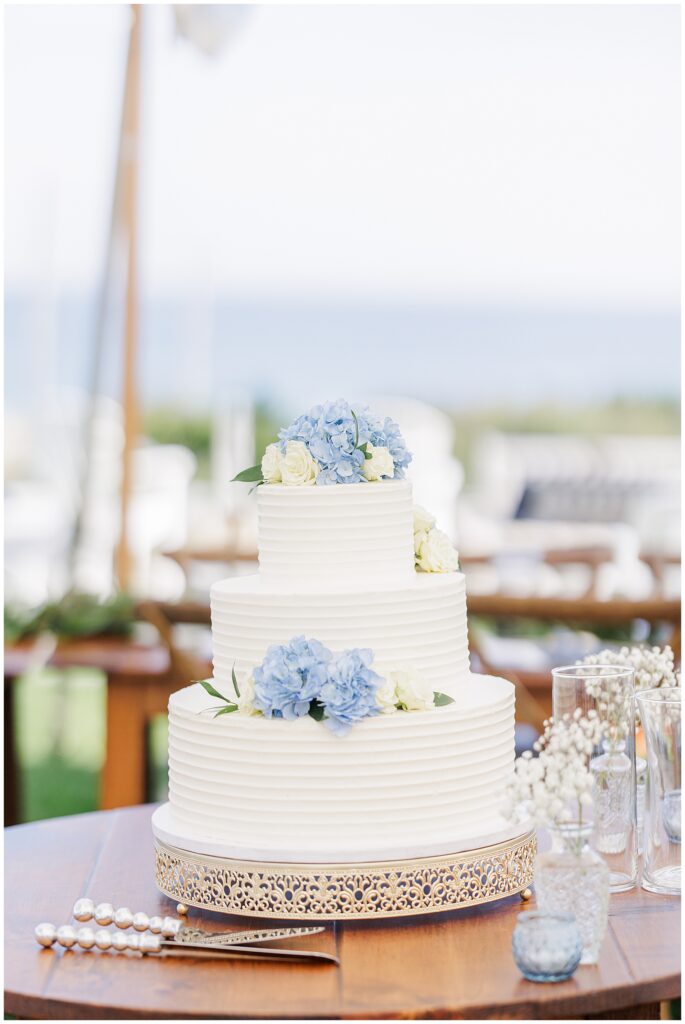 Three-tier white wedding cake decorated with blue and white flowers, displayed on a wooden table under a tent with ocean views at Nauset Beach Inn in Cape Cod.