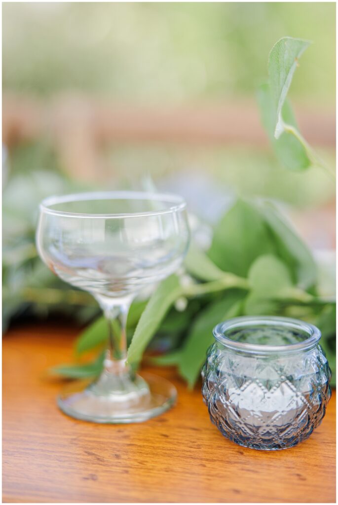 Close-up of a clear glass coupe and a small blue candle holder with greenery in the background, part of the elegant reception table decor at Nauset Beach Inn in Orleans, MA.