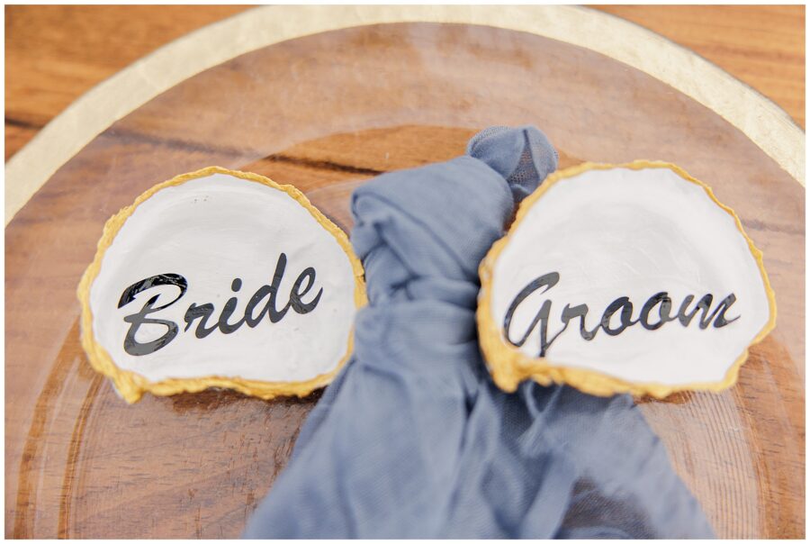 Place settings featuring oyster shell name cards reading “Bride” and “Groom,” styled with blue napkins on wooden plates at Nauset Beach Inn in Orleans, MA.