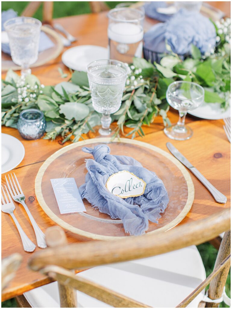 Close-up of a guest place setting with a gold-rimmed glass plate, blue napkin, and oyster shell name card reading “Colleen,” surrounded by greenery at Nauset Beach Inn in Cape Cod.