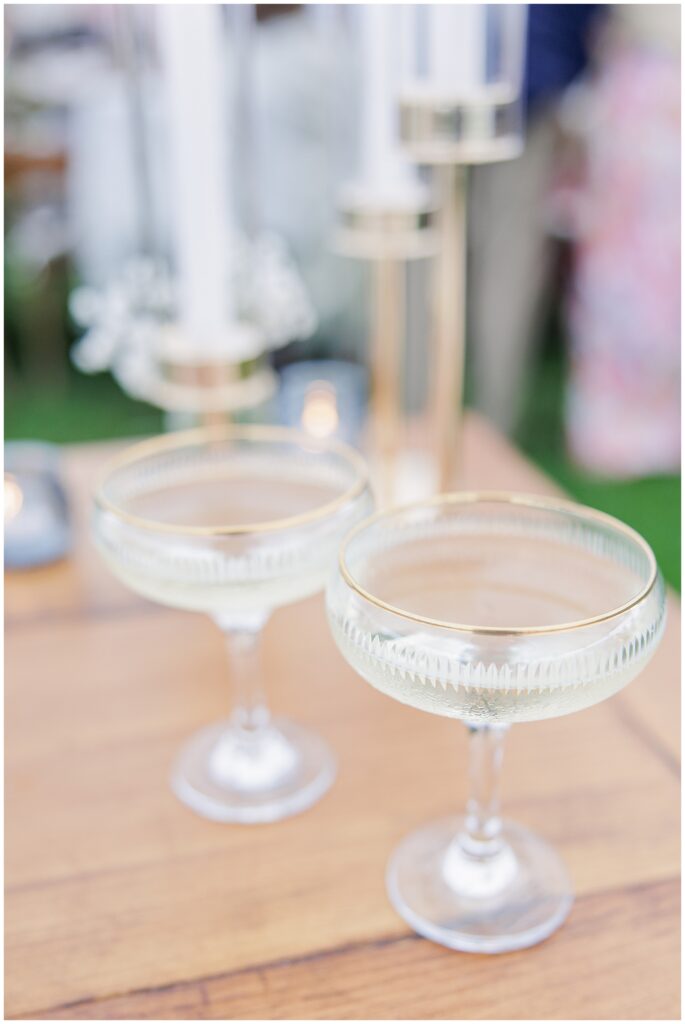Two champagne coupes filled with bubbly on a wooden table beside gold candle holders, part of the reception setup at Nauset Beach Inn in Cape Cod.