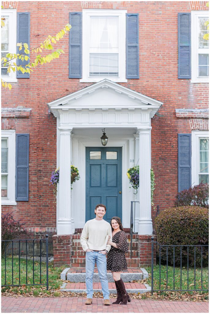 : A couple poses in front of a historic red brick home on a residential street in Newburyport, Massachusetts, during their engagement session.