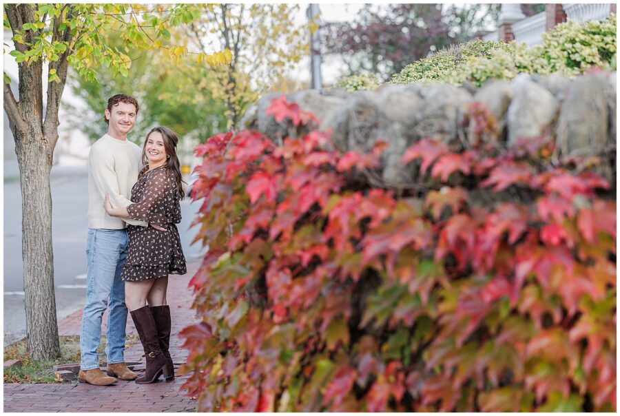 The couple smiles and embraces on a quiet sidewalk near Bartlet Mall in Newburyport, with red ivy on a stone wall and yellow-leaved trees in the background.